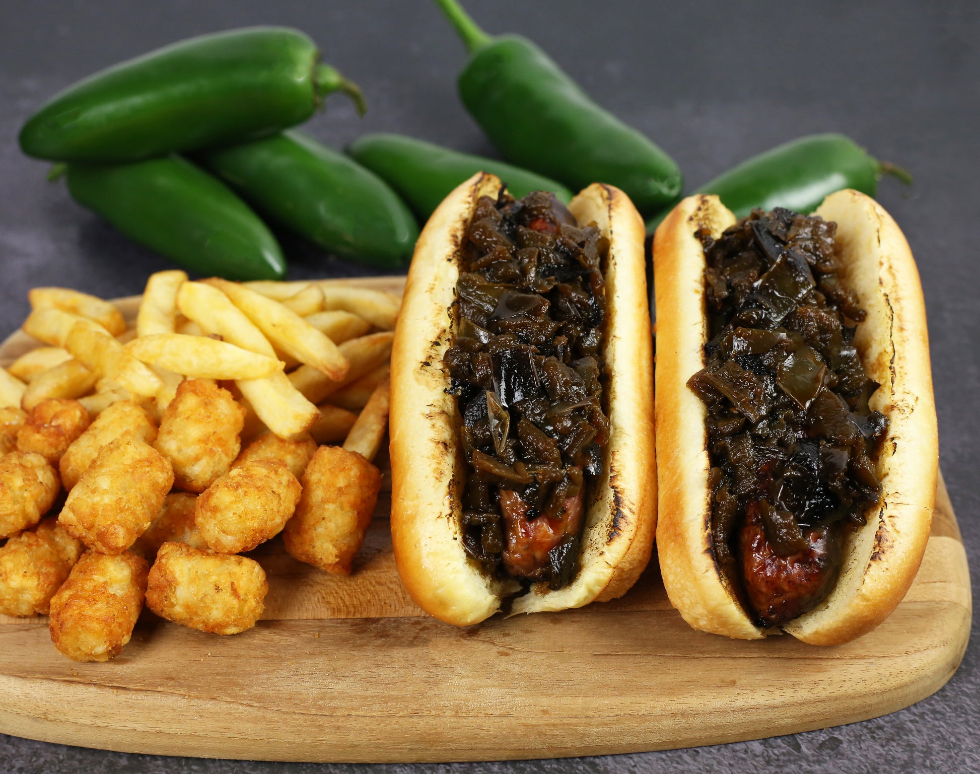 Two hot dogs with specialty jalapeño soy sauce on buns with tater tots, and fresh jalapeños on a wooden board.