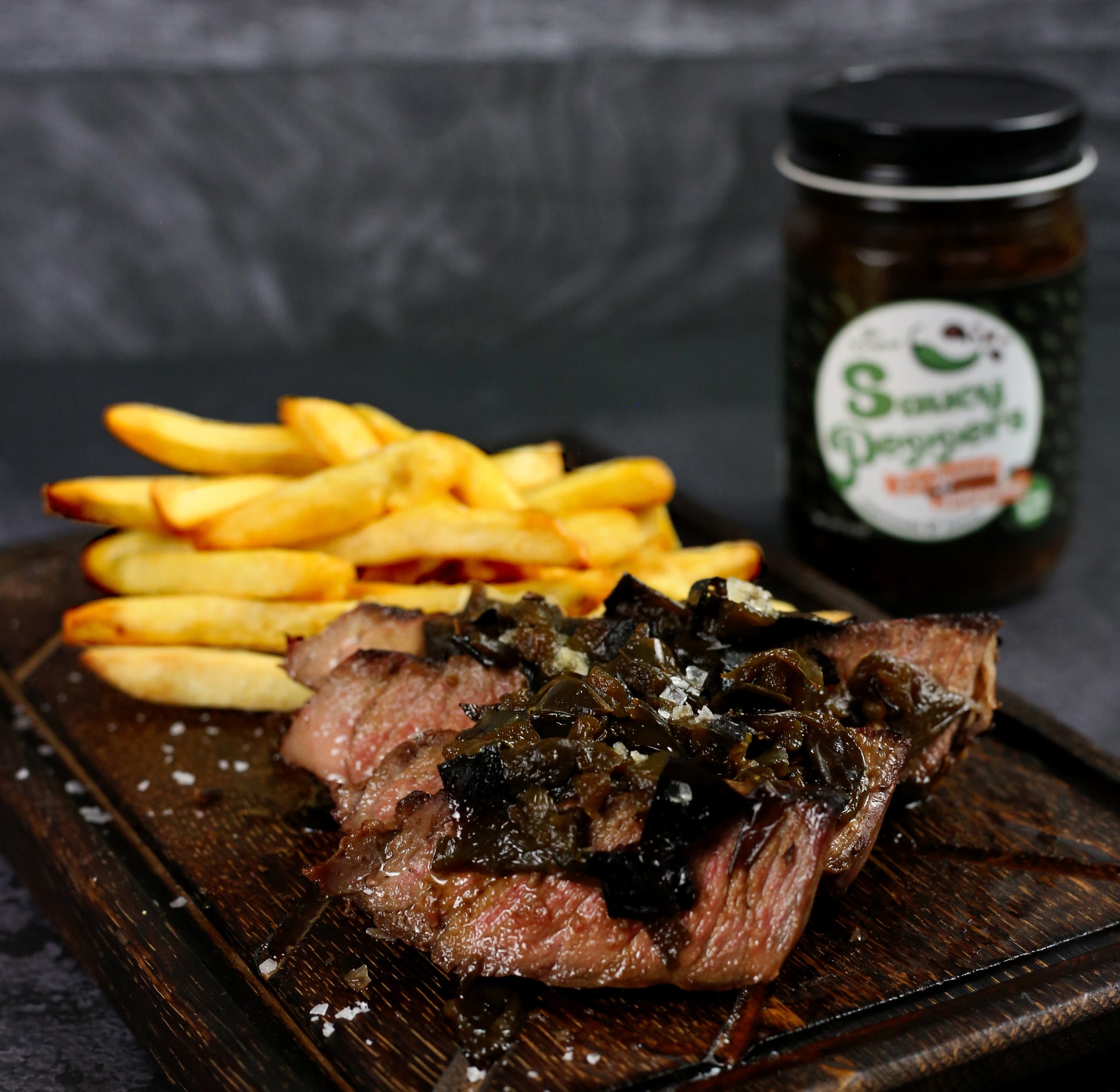 Steak with specialty  jalapeño soy sauce and fries on a wooden board with a jar of Saucy Peppers sauce in the background.