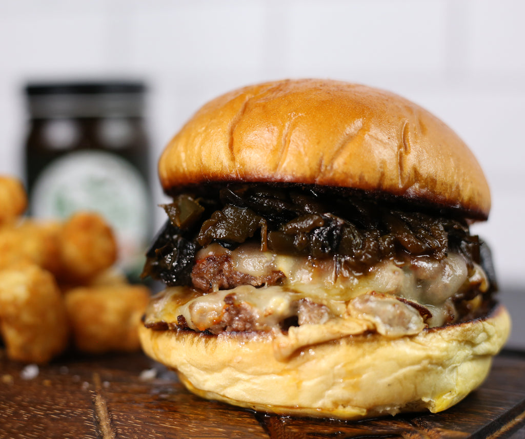 Deluxe burger with  jalapeño soy sauce, and cheese on a wood tray with tater tots in the background.