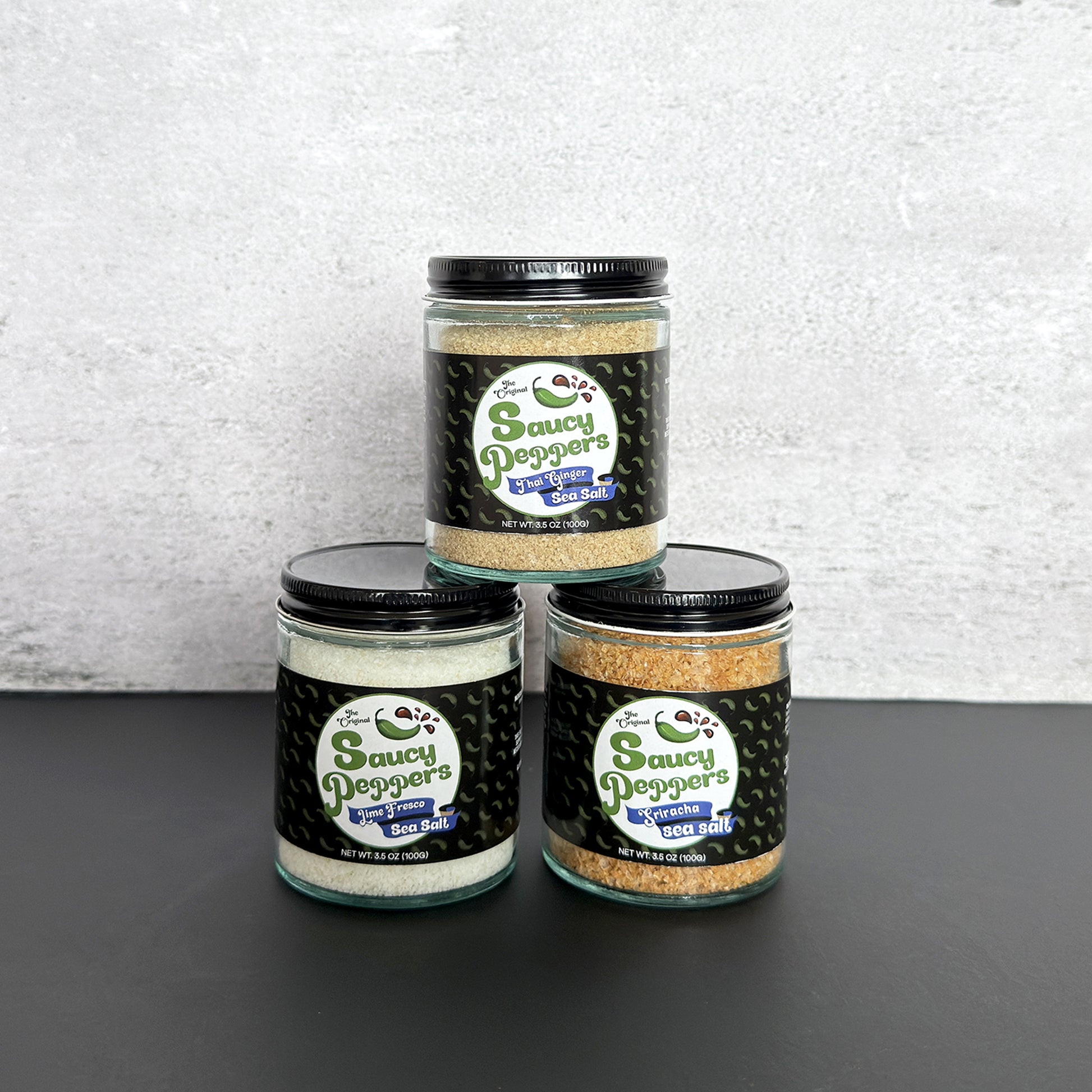 Three glass jars of infused sea salt seasoning blends arranged in a pyramid on a dark surface against a light gray textured wall. Flavored salts are Ginger, Lime and Sriracha